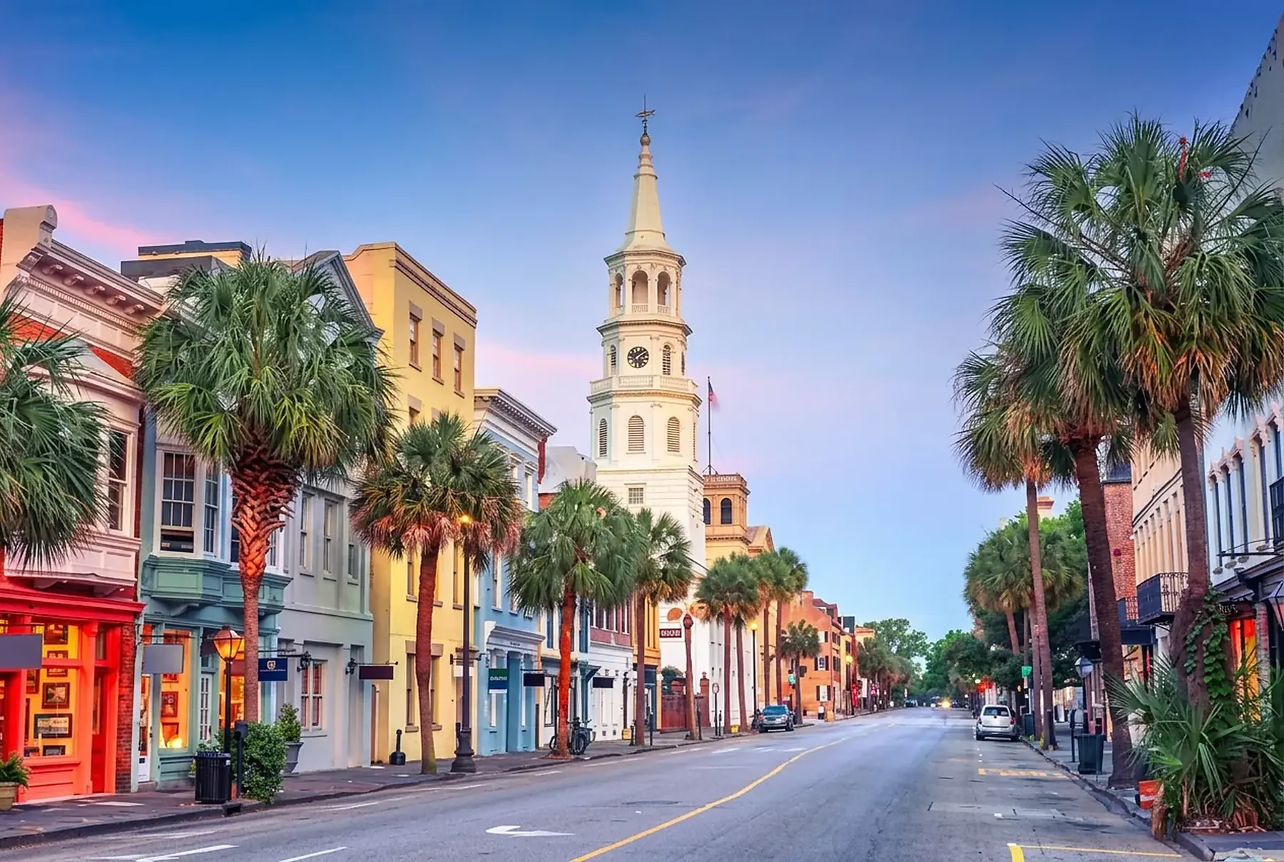 Palmetto lined street of historic Charleston storefronts and a white church steeple at dusk