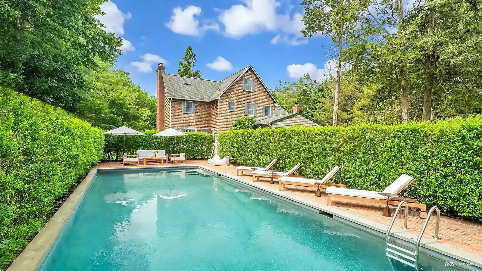 Cedar-shingle vacation home behind tall clipped hedges, with a long turquoise pool and teak sun loungers
